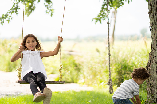 A sweet little girls is seen sitting on a swing as she glides back and forth in the warm summer air.  She is dressed casually and smiling as her little brother plays off to the side.