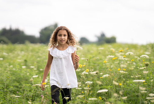 A sweet little curly haired girl walks through a field of white flowers on a sunny summer day.  She is dressed casually and has a smile on her face as she basks in the warm summer air and enjoys her time outdoors.