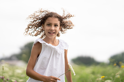 A sweet little curly haired girl runs through a field of white flowers on a sunny summer day.  She is dressed casually and has a smile on her face as she basks in the warm summer air and enjoys her time outdoors.