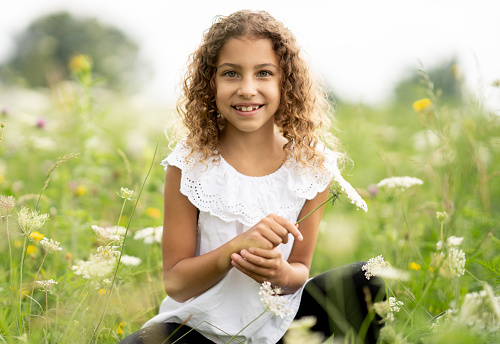 A sweet little curly haired girl sits cross-legged in a field of flowers as she basks in the warm summer air.  She is dressed casually and smiling as she picks a few to make a bouquet to take home.