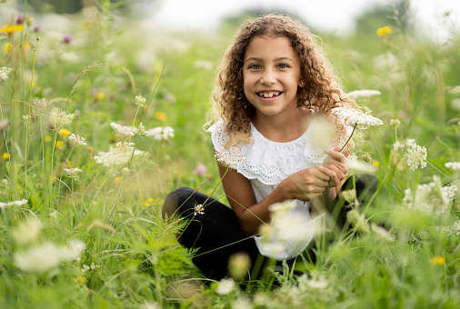 A sweet little curly haired girl sits cross-legged in a field of flowers as she basks in the warm summer air.  She is dressed casually and smiling as she picks a few to make a bouquet to take home.