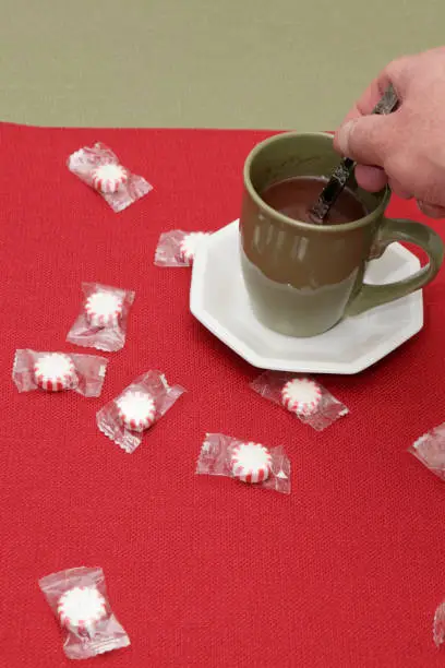 Adult caucasian hand stirring hot chocolate gourmet mix in a green cup with a handle near some individually wrapped peppermint candy. Peppermint candy near a male hand stirring hot chocolate drink.