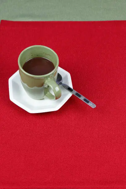One serving of hot chocolate gourmet mix in a coffee mug resting on a white octagon saucer on a red place mat with a green table cloth behind it. Liquid hot chocolate cocoa mix in a cup ready to drink