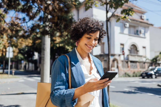 a delighted african-american woman texting on her smartphone while walking through the city - woman on phone stockfoto's en -beelden