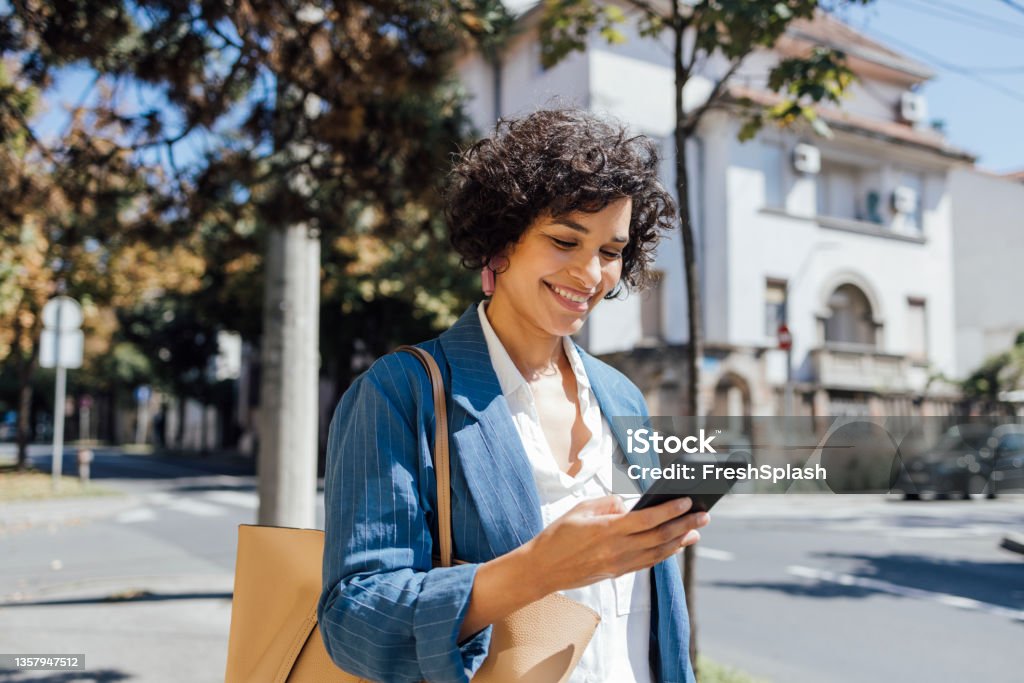 A Delighted African-American Woman Texting On Her Smartphone While Walking Through The City A mixed-race businesswoman reading something funny on her mobile phone on the street while going back home from work. People Stock Photo