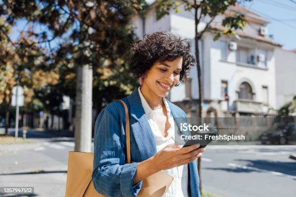 A Delighted Africanamerican Woman Texting On Her Smartphone While Walking Through The City Stock Photo - Download Image Now