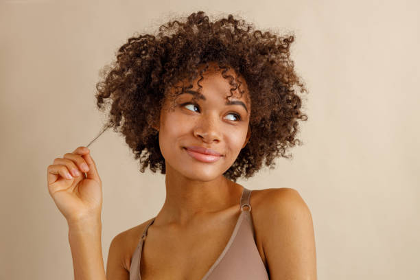 Charming multiracial woman posing against beige wall Cute multiracial woman, holding hair curl and looking sideways with flirtatious face expression, isolated on beige color background curly hair stock pictures, royalty-free photos & images