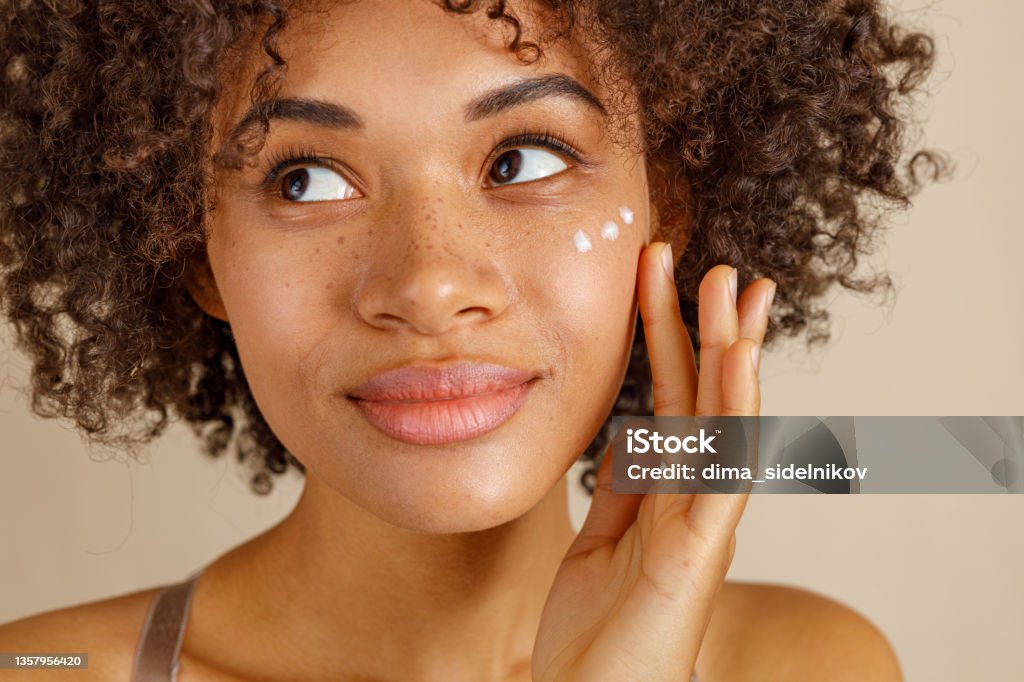 Cute lady using cosmetic product in studio Charming young lady looking at side with dots of face cream on her cheek, isolated on beige color background Skin Care Stock Photo