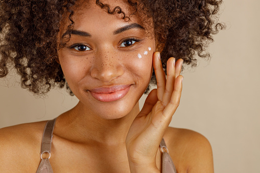 Happy multiethnic woman touching her clean smooth skin while applying day cream, isolated on beige color background