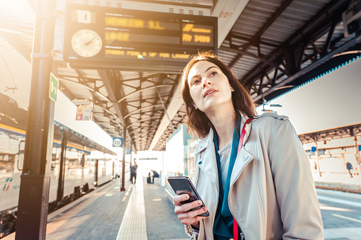 Young woman with departure times behind her waiting for her train while holding her mobile phone - Woman looking at the clock in the train station while her train is delayed - Transportation and urban life concept