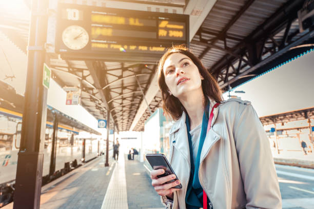 Young woman with departure times behind her waiting for her train while holding her mobile phone - Woman looking at the clock in the train station while her train is delayed - Transportation and urban life concept Young woman with departure times behind her waiting for her train while holding her mobile phone - Woman looking at the clock in the train station while her train is delayed - Transportation and urban life concept waiting telephone on the phone frustration stock pictures, royalty-free photos & images