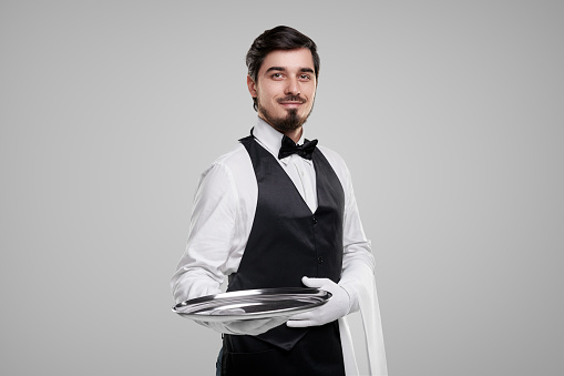 Positive young bearded waiter in elegant uniform and gloves carrying metal tray and looking at camera while representing restaurant service against gray background