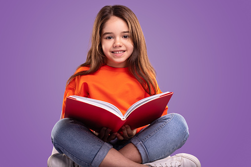 Happy child in casual clothes smiling and looking at camera while sitting crossed legged against violet background and reading book during school lesson