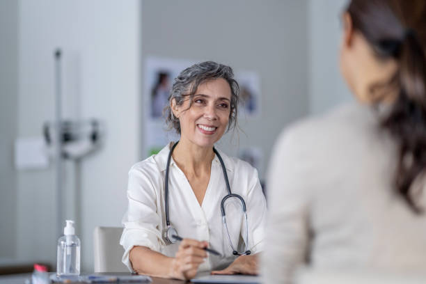 Doctor Sharing Test Results A mature female doctor points to the screen of her tablet laying out on the table in front of her as she shares her patients test results with her. She is wearing a white lab coat and has a stethoscope around her neck as she talks with the female patient of Asian decent. doctor general practitioner stock pictures, royalty-free photos & images