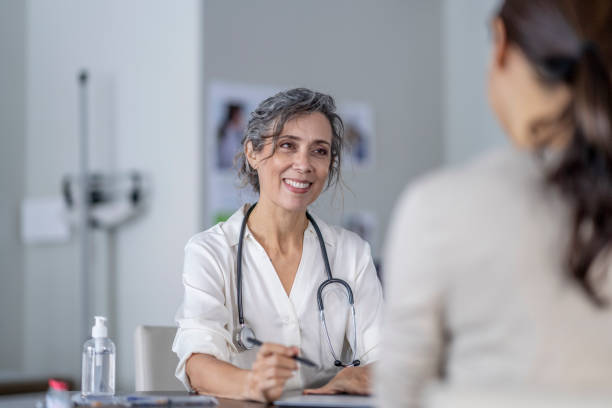 Doctor Sharing Test Results A mature female doctor points to the screen of her tablet laying out on the table in front of her as she shares her patients test results with her.  She is wearing a white lab coat and has a stethoscope around her neck as she talks with the female patient of Asian decent. general practitioner stock pictures, royalty-free photos & images