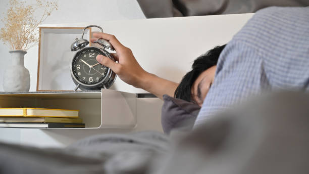 男はベッドに横たわって、目覚まし時計のスイッチをオフにするために手を伸ばします。 - oversleep ストックフォトと画像