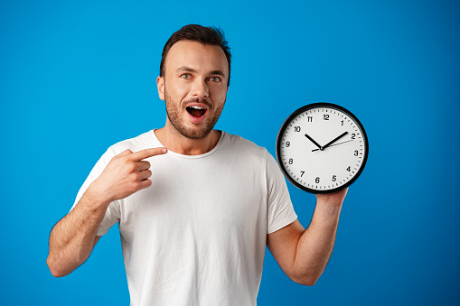 Handsome young man in white t-shirt posing with clock against blue background, close up