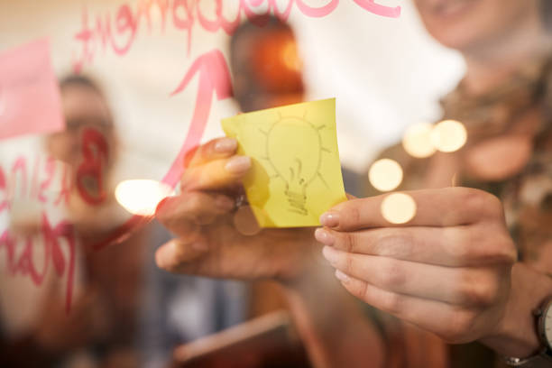 Close-up of creative woman applying sticky note with light bulb on a glass wall during business meeting in the office.