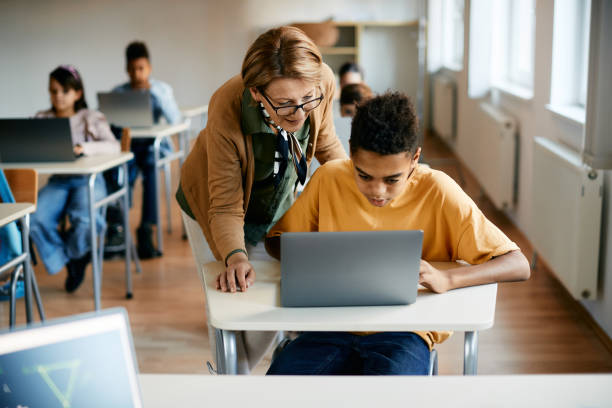 istock Black elementary student using laptop with help of a teacher in the classroom. 1358014273