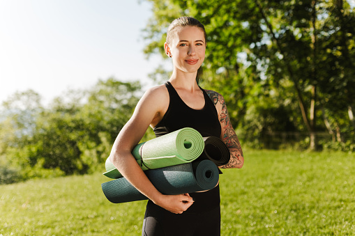 Portrait of smiling lady in black sporty top and leggings standing with yoga mats in hands and happily looking in camera with beautiful view on background