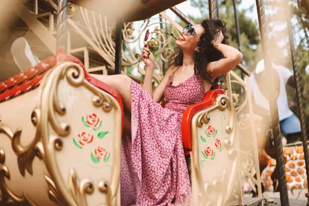 Prettygirl with dark curly hair in sunglasses and dress holding lolly pop candy in hand and happily looking aside while riding on carousel in amusement park