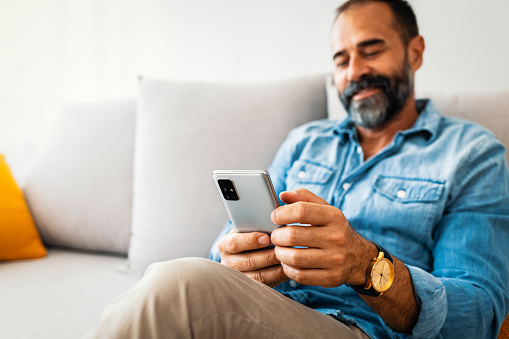 Man with spectacles relaxing sitting on couch while looking at mobile phone. Closeup of mature man using smartphone to checking email at home. Man reading email on smartphone