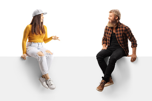 Casual young female sitting on a blank panel and talking to a bearded guy isolated on white background