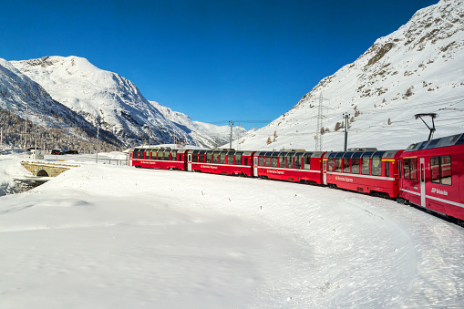 Grisons, Switzerland - November 05. 2021: The famous red Bernina Express tourist train is crossing the snow field in the Alps height.