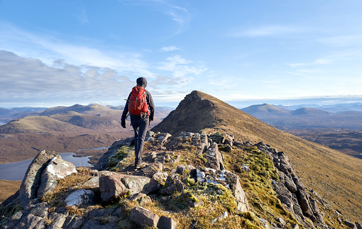 A hiker walking towards the summit of Sgurr an Tuill Bhain along a narrow rocky ridge in the Scottish Highlands on a sunny winters day.