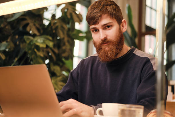 istock Young handsome bearded man intently looking in camera working on laptop in cafe 1358047779