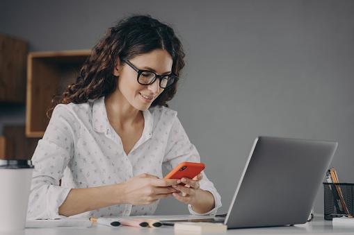 Young beautiful Italian businesswoman in glasses sits at desk in modern office at home and using modern smartphone, with pleasant smile looking at mobile phone and chatting in internet