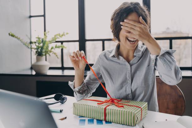 une jeune employée de bureau excitée fermant les yeux avec la main tout en déballant noël présente au travail - christmas office gift people photos et images de collection