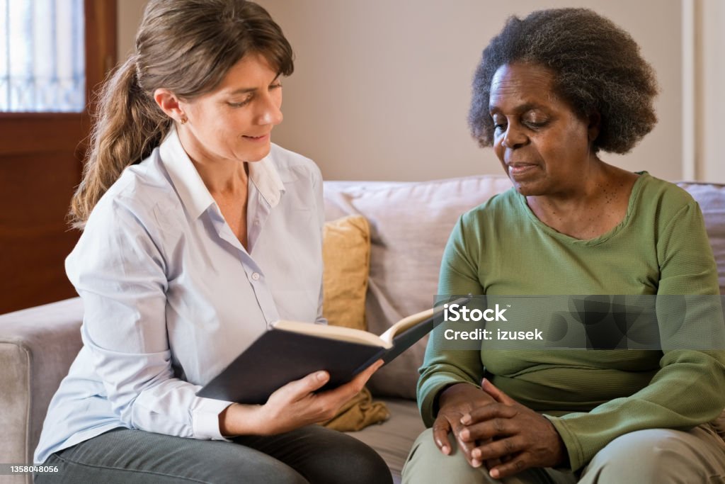 Female nurse reading book for senior woman at home Caregiver reading book for senior woman. Elderly female is sitting by nurse on sofa. They are in living room. 70-79 Years Stock Photo
