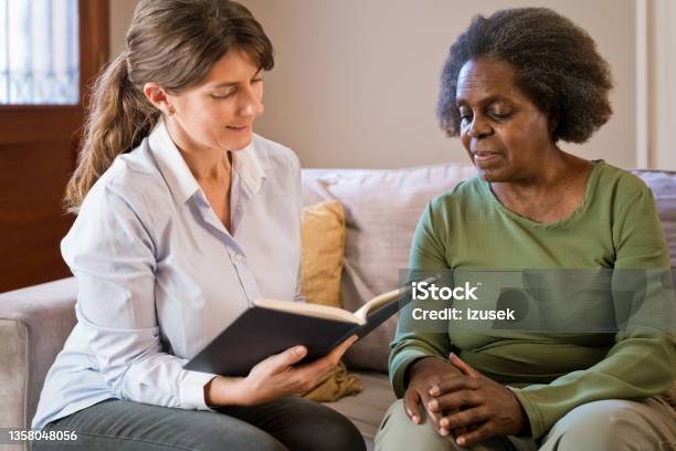 Female Nurse Reading Book For Senior Woman At Home Stock Photo - Download Image Now - 70-79 Years, Book, Community Outreach