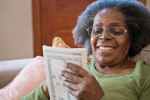 Smiling senior woman solving sudoku at home. Elderly female wearing eyeglasses sitting in living room. She is holding crossword puzzle book.