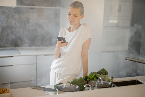 Caucasian apathetic woman watching something on smartphone before cooking dish from fresh vegetables and fruits. Concept of healthy and vegetarian eating. Kitchen in smoke at modern flat
