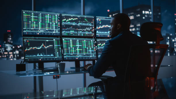 istock Hedge Fund Trader Working on a Computer with Multi-Monitor Workstation with Real-Time Stocks, Commodities and Exchange Market Charts. Businessman Works in Investment Bank Downtown Office at Night. 1358049223