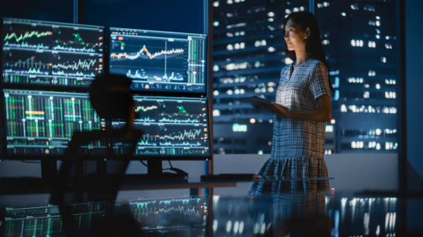 istock Financial Analyst Using Tablet Computer, Standing Next to Multi-Monitor Workstation with Real-Time Stocks, Commodities and Exchange Market Charts. Businesswoman Working in Investment Bank City Office. 1358049254