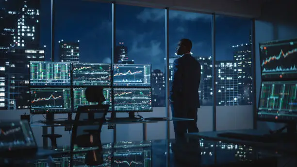 Photo of Successful Businessman Looking Out of the Window on Late Evening. Modern Hedge Fund Office with Computer with Multi-Monitor Workstation with Real-Time Stocks, Commodities and Exchange Market Charts.