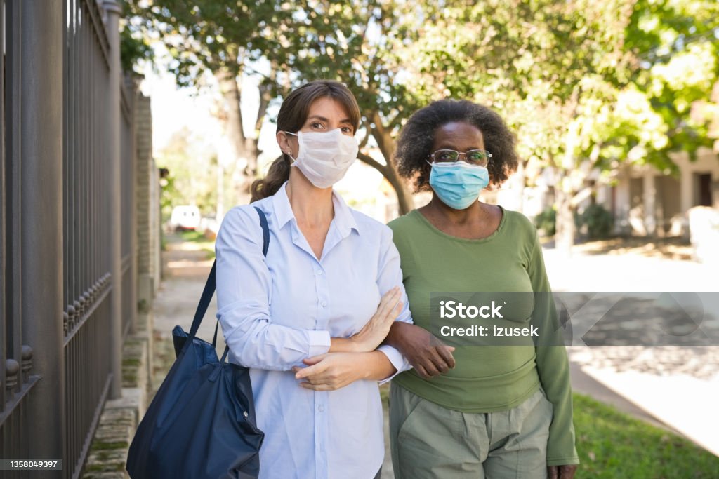 Nurse and senior woman walking on footpath during COVID-19 Portrait of caregiver and senior woman on footpath. Nurse and elderly female are walking with arm in arm. They are wearing surgical masks during COVID-19. Protective Face Mask Stock Photo