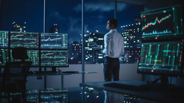 Financial Analyst Using Laptop Computer, Standing Next to Multi-Monitor Workstation with Real-Time Stocks, Commodities and Exchange Market Charts. Businessman Working in Broker Agency Office. analista financiero utilizando una computadora portátil, de pie junto a la estación de trabajo de múltiples monitores con acciones en tiempo real, materias primas y gráficos del mercado de divisas. empresario que trabaja en la oficina de la agencia de