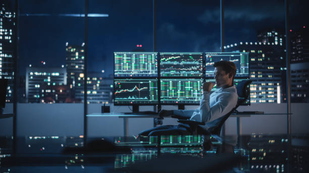 Financial Analyst Working on a Computer with Multi-Monitor Workstation with Real-Time Stocks, Commodities and Exchange Market Charts. Businessman Works in Investment Bank Downtown Office at Night. analista financiero que trabaja en una computadora con estación de trabajo de múltiples monitores con acciones en tiempo real, materias primas y gráficos del mercado de divisas. empresario trabaja en la oficina del centro del banco de inversión por la