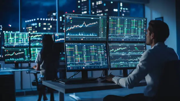 Photo of Financial Analyst Working on a Computer with Multi-Monitor Workstation with Real-Time Stocks, Commodities and Foreign Exchange Charts. Businessman Works in Investment Bank City Office at Night.