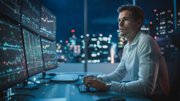 istock Financial Analyst Working on a Computer with Multi-Monitor Workstation with Real-Time Stocks, Commodities and Exchange Market Charts. Businessman Works in Investment Bank Downtown Office at Night. 1358050088