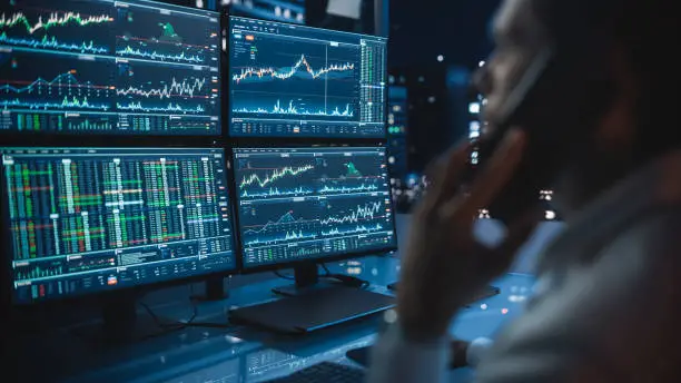 Photo of Financial Day Trader Working on a Computer with Multi-Monitor Workstation with Real-Time Stocks Charts and Talking on a Phone with a Client, While in Office Late in the Evening.