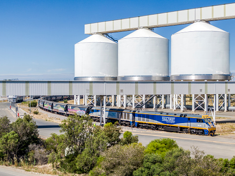Port Adelaide, Australia - Nov 25, 2021: Elevated view loaded ORA (One Rail Australia) grain train at Viterra Pelican Point unloading & storage facility. The huge storage bins tower over the train and road trucks.  Locomotives are on lease from Rail First Asset Management.