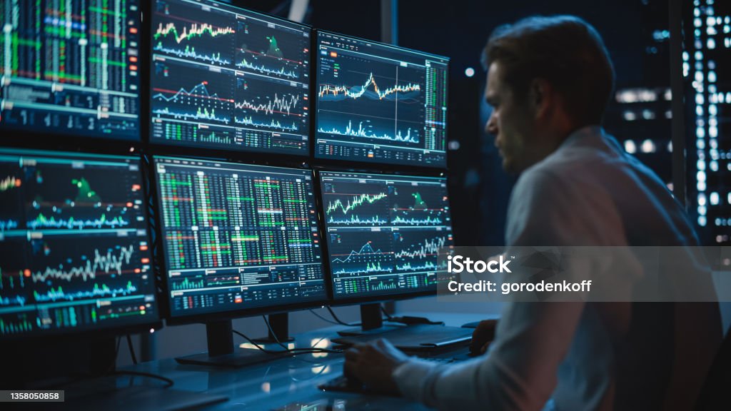 Financial Analyst Working on a Computer with Multi-Monitor Workstation with Real-Time Stocks, Commodities and Exchange Market Charts. Businessman Works in Investment Bank Downtown Office at Night. Stock Market and Exchange Stock Photo