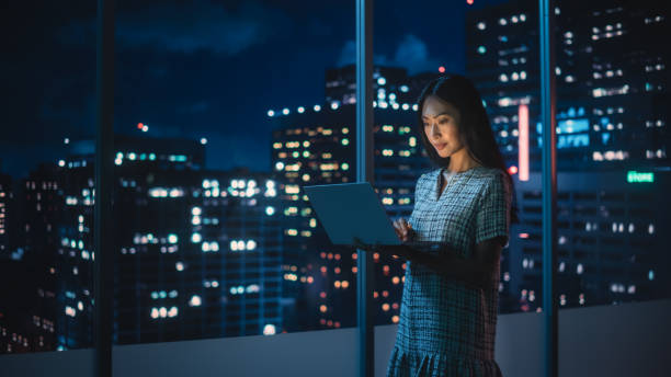 istock Financial Analyst Using Laptop Computer, Standing Next to Window with Beautiful Night City Skyline with Skyscrapers. Confident Asian Businesswoman Working in Modern Corporate Office. 1358050956