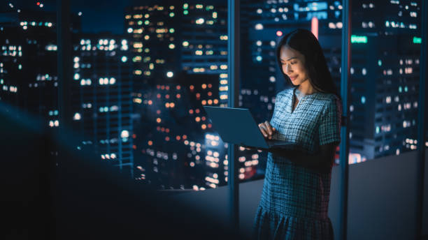 analista financiero usando computadora portátil, de pie junto a la ventana con hermoso horizonte nocturno de la ciudad con rascacielos. mujer de negocios asiática segura de sí misma que trabaja en una oficina corporativa moderna. - street directory fotografías e imágenes de stock