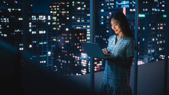 Financial Analyst Using Laptop Computer, Standing Next to Window with Beautiful Night City Skyline with Skyscrapers. Confident Asian Businesswoman Working in Modern Corporate Office.